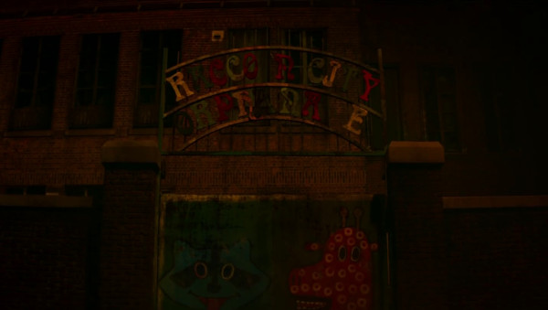 A still from Resident Evil: Welcome to Racoon City showing the gates to the Racoon City Orphanage. The gate has a friendly racoon (in teal) and giraffe painted on them, and the legend "Racoon City Orphanage" is shown above the gate, each letter is a different primary colours.