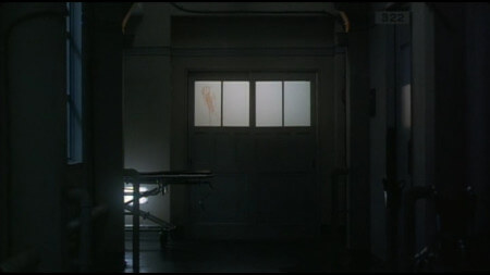 An ominous shot of a hospital corridor, with a set of double doors in the centre of the frame. The double doors have windows at about shoulder height, and one of the windows has a bloodied handprint smeared down it